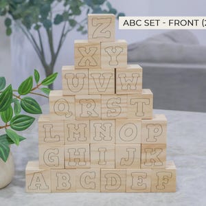 May include: A stack of 26 wooden alphabet blocks, each with a letter outlined on its face. The blocks are arranged in a pyramid shape, with the letters A through Z visible. The text "ABC SET - FRONT (26 PCS)" is at the top.