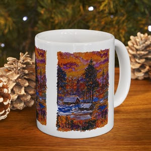 Puede incluir: Taza de cerámica blanca con una colorida pintura de paisaje de una cabaña en un bosque al atardecer. La obra de arte incluye árboles, un lago y un cielo vibrante. La taza tiene un asa curva y está sobre una superficie de madera.