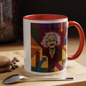 Puede incluir: Una taza de cerámica blanca con un borde y un asa rojos. La taza presenta un diseño abstracto colorido con un retrato de Albert Einstein en el centro. El diseño incluye flechas que apuntan en diferentes direcciones.