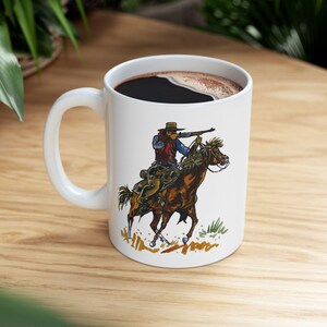 Puede incluir: Taza de cerámica blanca con una ilustración colorida de un vaquero montando un caballo y apuntando con un rifle. El vaquero lleva un sombrero marrón, una camisa azul y un chaleco marrón. El caballo es marrón con una crin y una cola blancas.