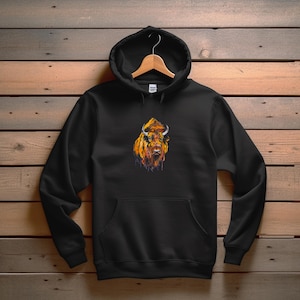 Puede incluir: Sudadera con capucha negra con un diseño de bisonte en acuarela. El bisonte está representado en tonos naranja, amarillo y marrón, con cuernos blancos. La sudadera tiene capucha, bolsillo delantero y está colgada de una percha de madera.