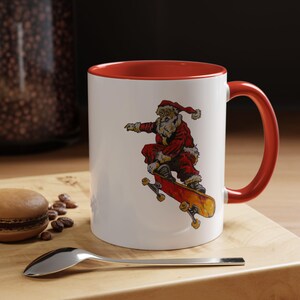 Puede incluir: Taza de cerámica blanca con interior y asa rojos. La taza presenta un gráfico de Papá Noel montando en monopatín sobre una patineta roja y amarilla. La taza está sobre una superficie de madera con una cuchara y un pastel.
