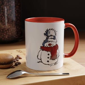 Puede incluir: Taza de cerámica blanca con interior y asa rojos. La taza presenta una ilustración caprichosa de un muñeco de nieve con un gorro azul y blanco y una bufanda roja. Una cuchara y un macaron están sobre una superficie de madera.