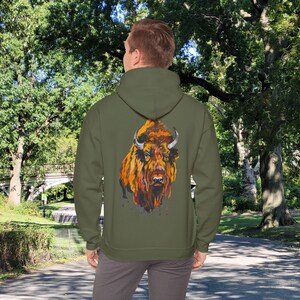 Puede incluir: Una sudadera con capucha verde con una ilustración colorida de un bisonte en la espalda. El bisonte está pintado en tonos de naranja, amarillo y marrón.