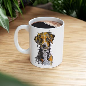 Puede incluir: Taza de café de cerámica blanca con asa negra. La taza tiene una ilustración colorida de un perro con pelaje marrón, negro y blanco. El perro está mirando directamente al espectador.