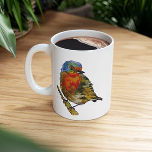 Puede incluir: Taza de café de cerámica blanca con una ilustración de acuarela de un pájaro colorido posado en una rama. El pájaro tiene plumaje azul, naranja, rojo y amarillo. La taza está llena de café oscuro y tiene un asa blanca.