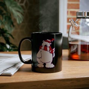 Puede incluir: Una taza de cerámica negra con un oso polar de dibujos animados que lleva una bufanda y un gorro a rayas rojas y blancas. La taza está sobre una superficie de madera, con vapor saliendo. Una tetera de cristal está en el fondo.