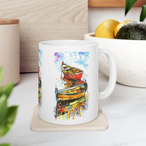 Puede incluir: Taza de cerámica blanca con una pintura acuarela colorida de dos barcos de madera sobre un fondo azul y blanco.