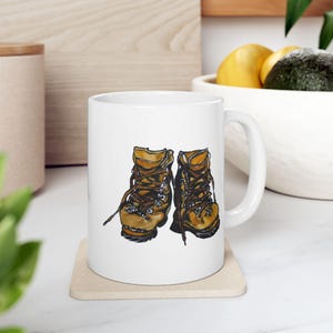 Puede incluir: Taza de cerámica blanca con una ilustración de dos botas de senderismo marrones y amarillas. Las botas tienen cordones y parecen gastadas.