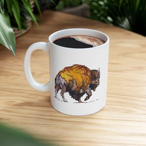 Puede incluir: Taza de café de cerámica blanca con una ilustración detallada de un bisonte. El bisonte tiene pelaje marrón, amarillo y negro. La taza está llena de café oscuro. La taza tiene un asa blanca y está sobre una superficie de madera.