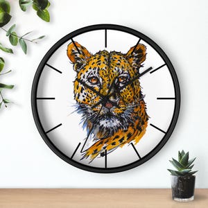May include: A round wall clock with a black frame featuring a watercolor illustration of a leopard's face. The leopard has orange and black spots, with a white background and black clock hands.