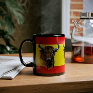 Puede incluir: Taza de cerámica negra con un diseño rojo y amarillo que presenta una cabeza de toro. El toro es marrón y negro con la nariz blanca y los cuernos.