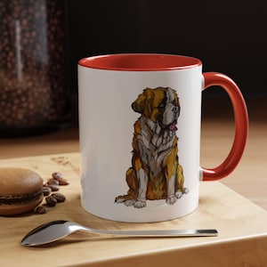 Puede incluir: Taza de cerámica blanca con interior y asa rojos. La taza presenta una ilustración detallada de un perro San Bernardo en tonos marrón, beige y blanco. La taza está sobre una superficie de madera.