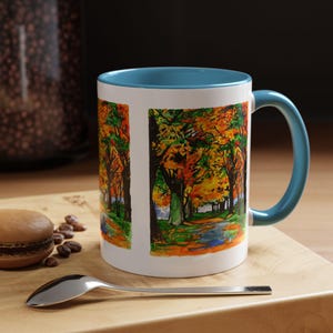 Puede incluir: Una taza de cerámica blanca con un borde y un asa de color azul verdoso. La taza presenta una ilustración colorida de un camino a través de un bosque con árboles en tonos de naranja, amarillo, verde y marrón.