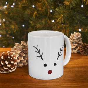 Puede incluir: Taza de cerámica blanca con un diseño de cara de reno. El diseño presenta cuernos negros, puntos negros para los ojos y un punto rojo para la nariz. La taza está sobre una superficie de madera con piñas y un fondo verde borroso.