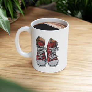 Puede incluir: Taza de cerámica blanca con un par de zapatillas rojas con cordones grises. La taza está llena de café oscuro. Las zapatillas son una ilustración en acuarela. La taza tiene un asa blanca.