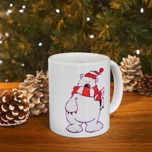 Puede incluir: Taza de cerámica blanca con un oso polar de dibujos animados que lleva una bufanda y un gorro a rayas rojas y blancas. El oso está delineado en morado. La taza tiene un asa blanca. Fondo de hojas de pino y piñas.