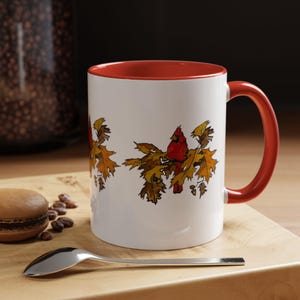 Puede incluir: Taza de cerámica blanca con borde y asa rojos. La taza presenta una ilustración colorida de un cardenal posado en una rama con hojas de otoño.