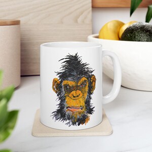 Puede incluir: Taza de cerámica blanca con una ilustración en negro y amarillo de un chimpancé con un mohawk. El chimpancé tiene una expresión seria.