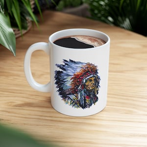 Puede incluir: Taza de café de cerámica blanca con una colorida ilustración de un jefe nativo americano con un tocado de plumas. La taza está llena de café oscuro y tiene un asa blanca. El diseño es de estilo acuarela.