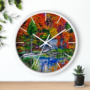 Puede incluir: Un reloj redondo blanco con una pintura de acuarela de una escena de lago con una casa y árboles en colores otoñales. La esfera del reloj tiene agujas negras y números negros.