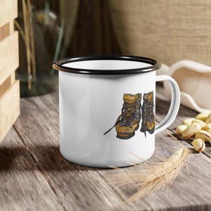 Puede incluir: Taza de esmalte blanco con borde negro que presenta una ilustración dibujada a mano de dos botas de senderismo marrones con cordones.