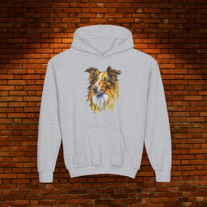 Puede incluir: Una sudadera con capucha gris claro con una ilustración en acuarela de la cabeza de un perro Collie. El pelaje del perro está representado en tonos marrones, dorados y blancos, con una textura esponjosa. La sudadera tiene una capucha con cordón y un bolsillo tipo canguro.