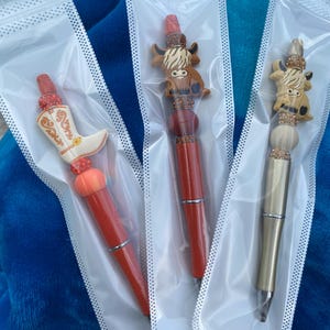 May include: Three pens with different animal toppers. The first pen has a red and orange boot topper with rhinestones. The second pen has a brown and white cow topper with rhinestones. The third pen has a gold and brown cow topper with rhinestones.
