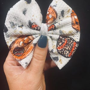 May include: A white fabric hair bow with a baseball glove and leopard print design. The bow features orange, black, and gray accents. The bow is held in a hand against a black background.