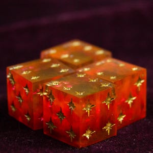 Op de afbeelding: Vier doorschijnende oranje dobbelstenen met gouden sterinsluitsels. De dobbelstenen zijn in een cluster gerangschikt en tonen de ingebedde sterontwerpen. De dobbelstenen staan tegen een donkerpaarse achtergrond, wat de levendige kleuren en details benadrukt.