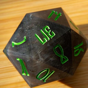 May include: A large, multi-sided die with a dark, marbled appearance. The numbers and the word "LIE" are etched in bright green. The die is designed for tabletop gaming.