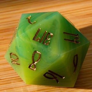 May include: A large, multi-sided die with a green and yellow marbled design. The word "LIE" is visible in gold lettering. Other sides feature gold numerals. The die is designed for tabletop role-playing games.