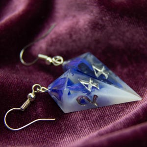 Può includere: Un paio di orecchini fatti a mano con un design geometrico. Gli orecchini presentano una resina trasparente con accenti blu e bianchi e ganci in metallo color argento. Gli orecchini hanno il numero 4.