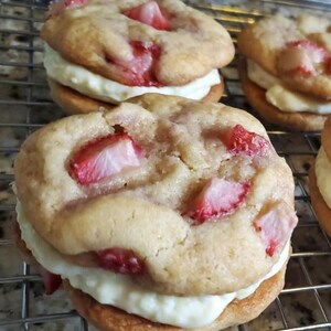 May include: Close-up of three strawberry shortcake cookies. Each cookie is made with a light brown cookie, a layer of white frosting, and fresh strawberries.