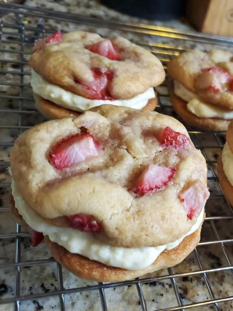 May include: Close-up of three strawberry shortcake cookies. Each cookie is made with a light brown cookie, a layer of white frosting, and fresh strawberries.