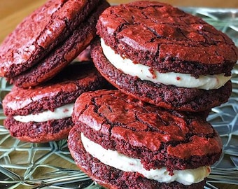Puede incluir: Cuatro galletas de terciopelo rojo con glaseado blanco, apiladas en un plato de vidrio transparente. Las galletas son suaves y tienen una textura desmenuzable.