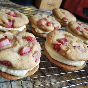 May include: A close-up of a wire rack with six homemade strawberry shortcake cookies. The cookies are filled with a white cream and topped with fresh strawberries.