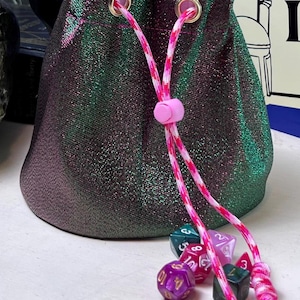 May include: A drawstring pouch made of shimmering, color-shifting fabric that transitions between deep green and dark purple. The bag features gold-toned metal grommets and a pink and white patterned cord with a pink plastic toggle and knotted ends.