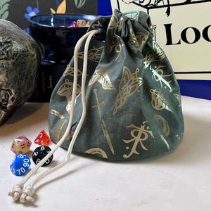 May include: A metallic silver skull, a dark green drawstring pouch with gold fantasy-themed symbols, and a blue sign featuring a treasure chest illustration and the text The Luddy's Loot, accompanied by several small multi-colored polyhedral dice.
