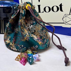 May include: A dark green drawstring pouch featuring a gold and multi-colored floral pattern, accented with a silver-toned metal charm. A collection of multi-colored, multi-faceted polyhedral dice with white numbers are arranged in front of the pouch.