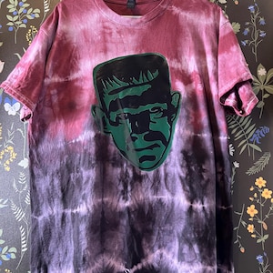 May include: A tie-dye t-shirt with a gradient of burgundy, purple, and black. The shirt features a green graphic of Frankenstein's monster on the front. The shirt is hanging on a white hanger.