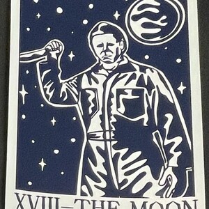 May include: A navy blue and white sticker featuring a stylized illustration of a figure holding a knife, set against a starry night sky with a moon. The text "XVIII-THE MOON" is at the bottom.