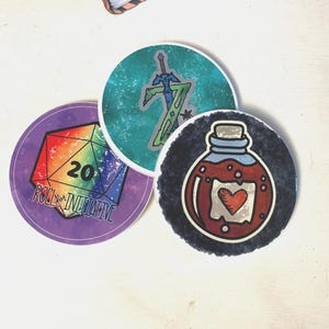Puede incluir: Tres pegatinas circulares con diseños de fantasía. Una muestra un dado de 20 caras arcoíris con el texto Roll for Initiative. Otra muestra una espada sobre un fondo verde azulado. La tercera representa una botella de poción roja con un corazón.