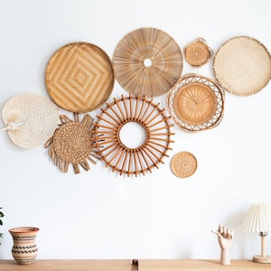 Puede incluir: Una colección de decoraciones de pared tejidas en varias formas y tamaños, incluyendo diseños redondos, en forma de abanico y de sol. Los tonos beige y marrón naturales crean una estética cálida y texturizada. La disposición se muestra contra una pared blanca.