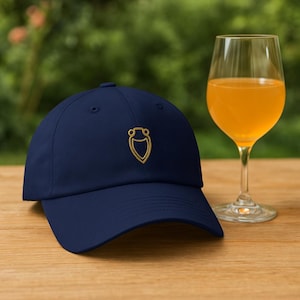 Pode incluir: Um boné de beisebol azul marinho com um design bordado em dourado de um escudo estilizado com um círculo no topo.