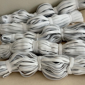 May include: Several bundles of white elastic cord are neatly tied with matching elastic. The cords are tightly wound and appear to be ready for use in various sewing or crafting projects. The neutral color makes them versatile.