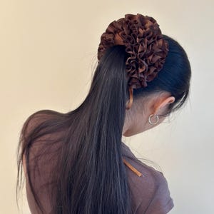 May include: A brown hair accessory with a ruffled flower design. The hair piece is attached to a ponytail. The hair is long and dark brown. The person is wearing a brown top and a silver hoop earring.