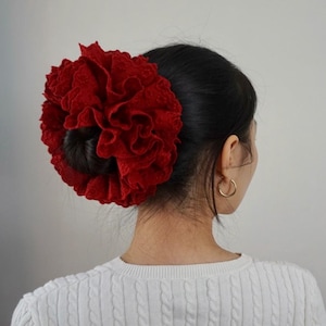 Peut inclure: Un grand accessoire pour cheveux rouge et froissé, ressemblant à une fleur, est porté en chignon. La coiffure est le point central, avec un aspect texturé. La personne porte un pull blanc et des créoles dorées.