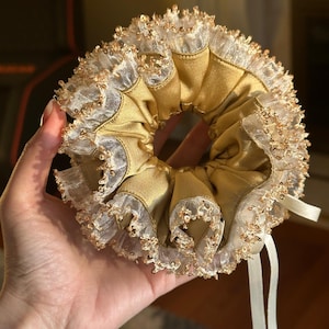 Peut inclure: Un chouchou en satin doré avec une bordure en dentelle blanche et des paillettes dorées. L'accessoire pour cheveux a un design à volants et un ruban blanc. Le chouchou est tenu dans une main.