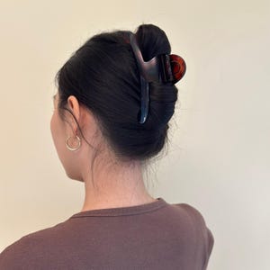 May include: A dark brown and tortoiseshell hair claw clip securing a bun. The clip is made of a glossy material and has a curved design. The person is wearing a brown shirt and gold hoop earrings.